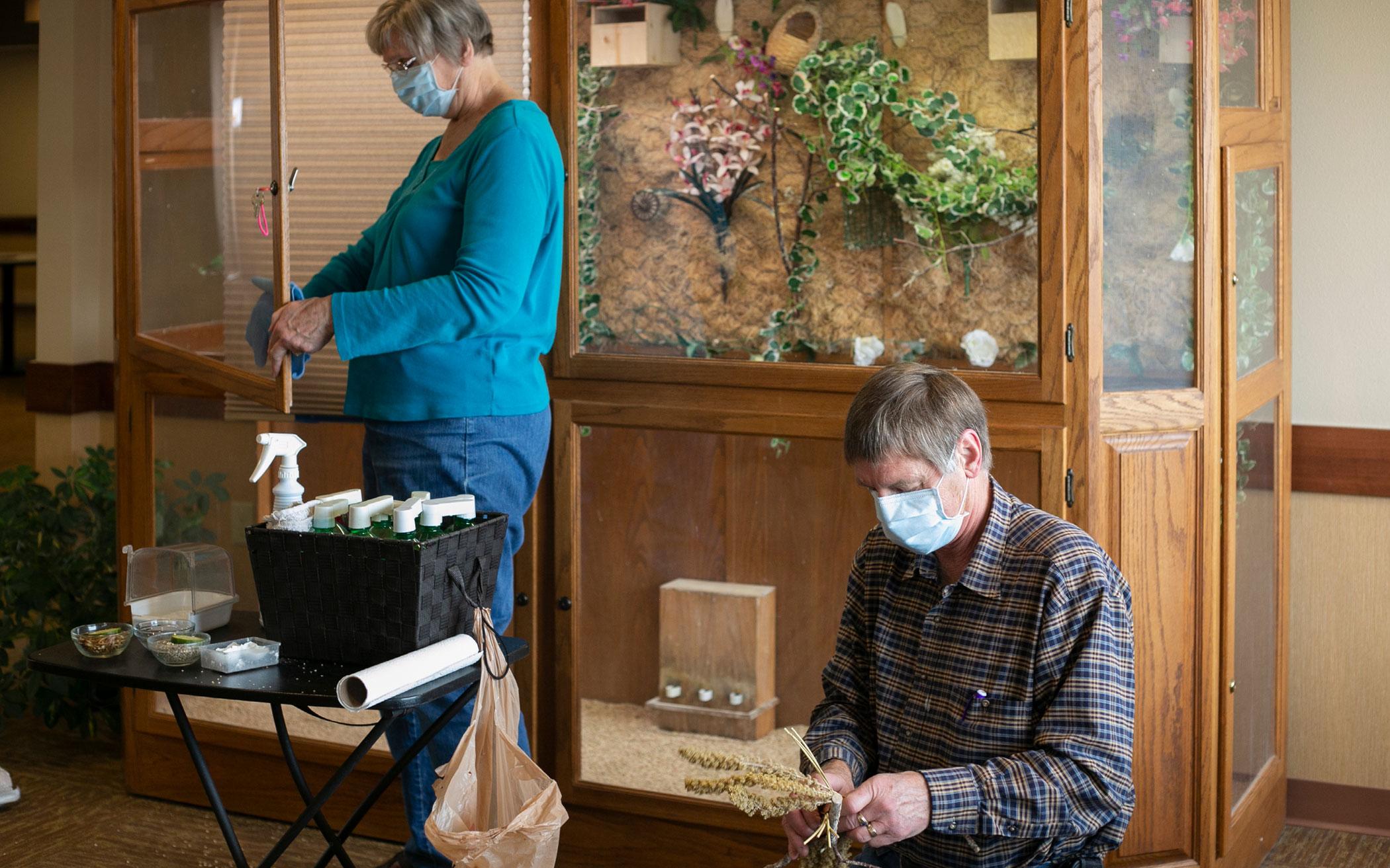 Mary Elgersma (left) cleans a window in a Pine Haven aviary while Mel removes old millet sprays from a wooden perch and replaces them with fresh millet, a food source.